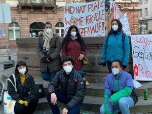 Das Antirassismus Referat blickt in die Kamera. Sie tragen Masken und stehen vor Bannern, um zu protestieren.